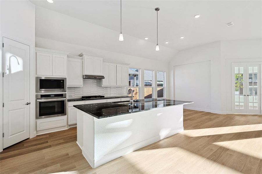 Kitchen with vaulted ceiling, appliances with stainless steel finishes, a kitchen island with sink, light wood-type flooring, and white cabinets