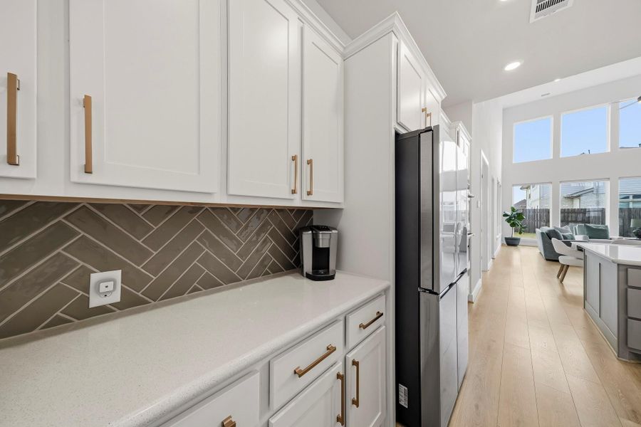A beautifully appointed coffee/serving bar elevates the kitchen with Quartz countertops, generous upper and lower cabinetry and the feel of a custom-designed entertaining feature. Perfect for crafting morning espresso, serving cocktails or displaying favorite glassware, it adds both sophistication and everyday function. The walk-in pantry sits just across for effortless access, while the refrigerator remains with the home for added move-in convenience.