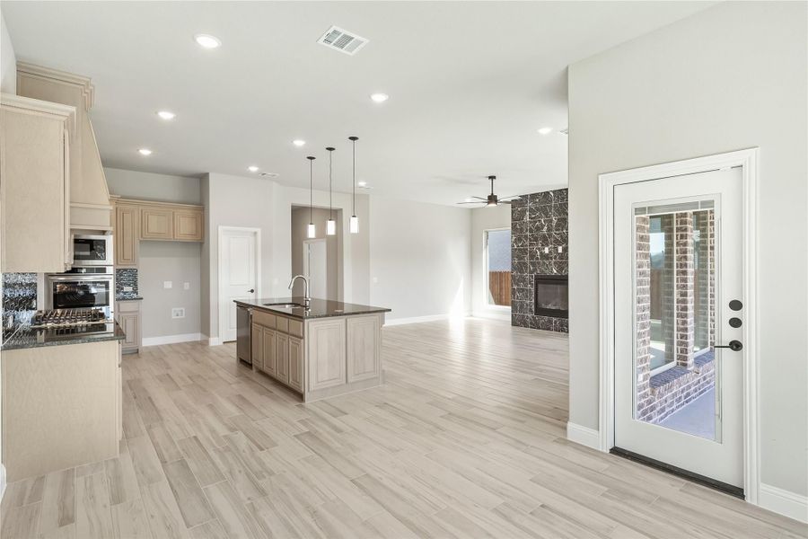 Open-concept kitchen and living area with center island, light wood floors, and stone fireplace visible in background. Open-concept kitchen and living area with center island, light wood floors, and stone fireplace visible in background.