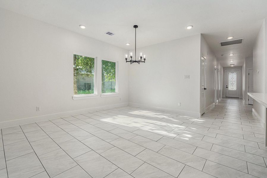 Bright and spacious room with large tiled flooring, neutral walls, and ample natural light from two windows. Features a modern chandelier and leads to a hallway with a decorative glass door. Perfect for a dining or living area.
