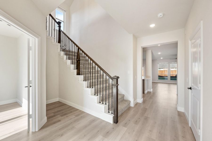 Entryway with carpeted staircase, dark iron balusters, and light hardwood floors leading to open living area Entryway with carpeted staircase, dark iron balusters, and light hardwood floors leading to open living area