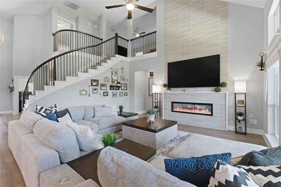Expansive living area featuring a two-story ceiling, a curved staircase with dark iron balusters, wood-finish flooring, and a tiled fireplace with an integrated linear electric insert