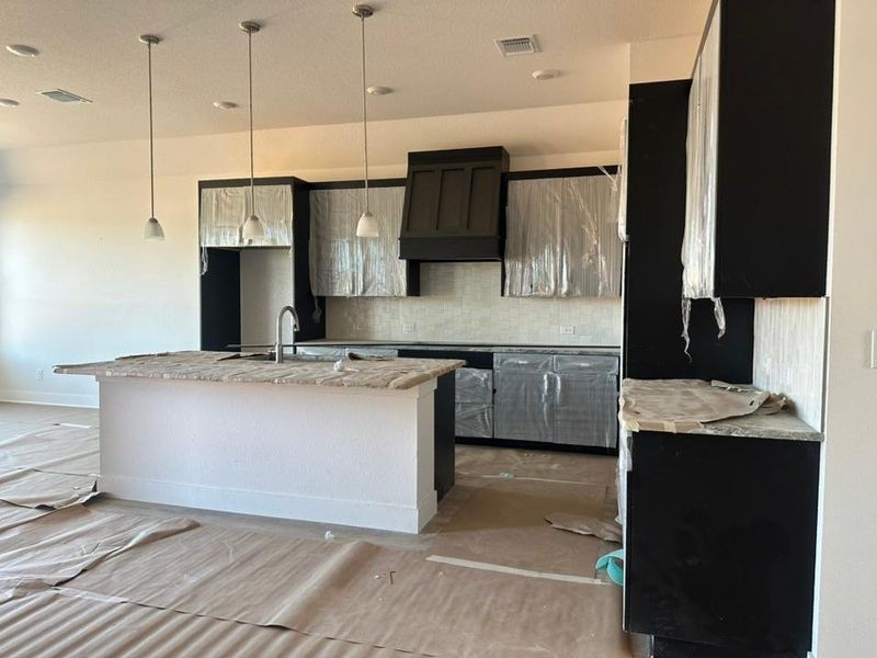 Kitchen featuring backsplash, hanging light fixtures, dark cabinetry, a kitchen island with sink, and custom range hood