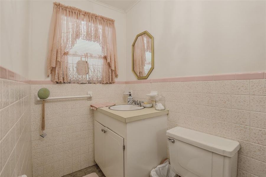 Bathroom featuring a vanity with an integrated sink, a toilet, an octagonal mirror with a gold-tone frame, and tiled walls with a pink accent stripe Bathroom featuring a vanity with an integrated sink, a toilet, an octagonal mirror with a gold-tone frame, and tiled walls with a pink accent stripe