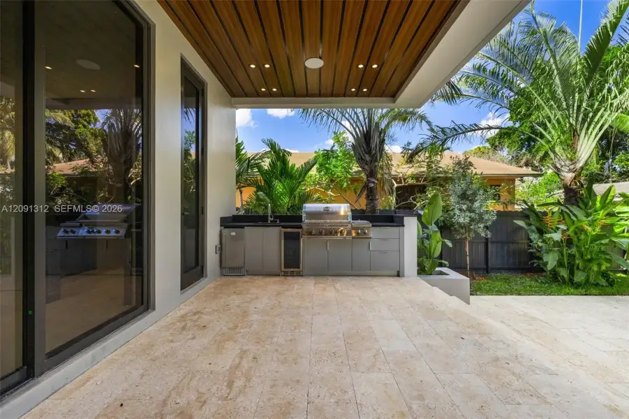 Covered outdoor kitchen featuring built-in cabinetry, stainless steel grill, beverage cooler, stone-finish flooring, and a wood-slat ceiling Covered outdoor kitchen featuring built-in cabinetry, stainless steel grill, beverage cooler, stone-finish flooring, and a wood-slat ceiling