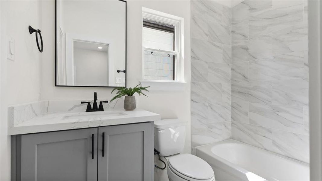 Modern secondary bathroom featuring a faux marble tile, a modern sleek gray custom vanity with a quartz countertop, custom modern framed matte black mirror, and a full-size bathtub surrounded by stone tiles.