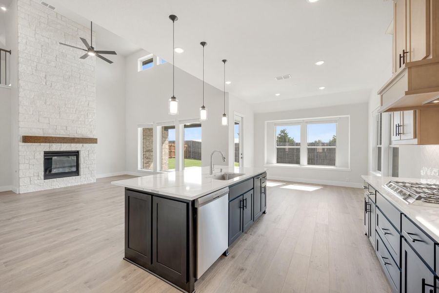 Open kitchen with dark island, quartz countertops, and stone fireplace in adjoining living area with vaulted ceiling Open kitchen with dark island, quartz countertops, and stone fireplace in adjoining living area with vaulted ceiling