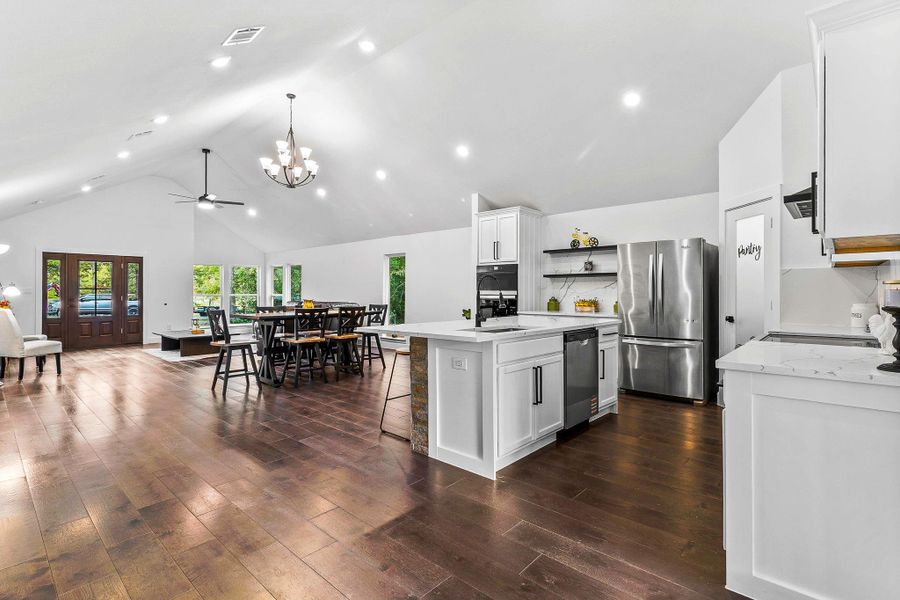 This photo showcases a spacious open-concept living area with a modern kitchen featuring white cabinetry, stainless steel appliances, and an island. The dining area offers ample seating, and the space is accented by high ceilings and large windows, creating a bright and inviting atmosphere.
