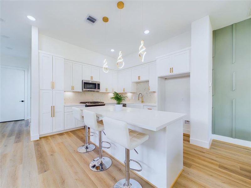 This modern open-plan kitchen features white cabinetry with gold color hardware, a farmhouse sink, central island with bar seating, sleek Stainless Steel appliances, and unique hanging light fixtures. The space is bright with natural light!