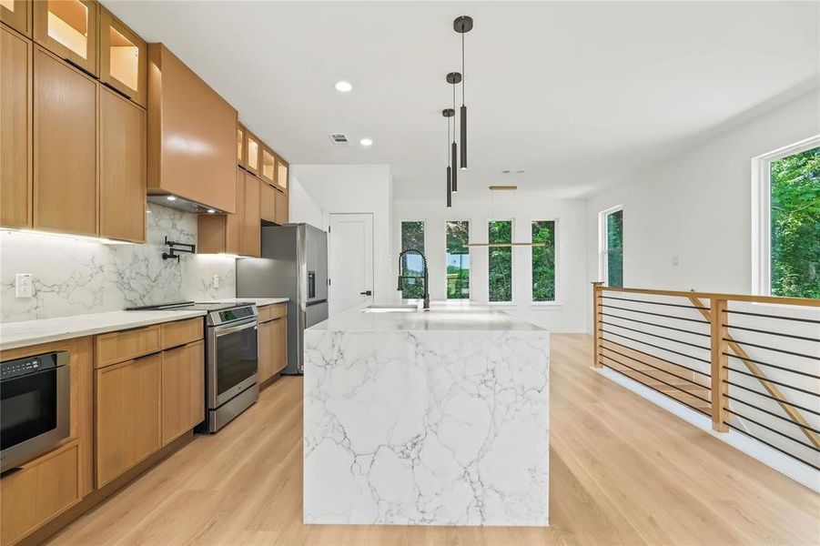 Kitchen featuring glass insert cabinets, appliances with stainless steel finishes, light stone countertops, a center island with sink, and decorative light fixtures