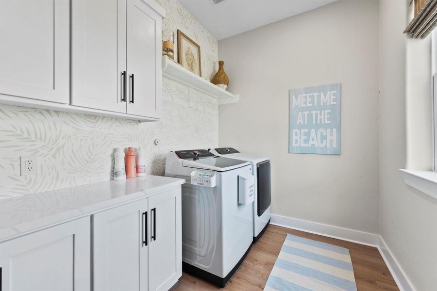 This laundry room features modern appliances, white cabinetry, and a decorative Serena & Lily accent wall. It offers ample counter space and shelving for storage, with a beach-themed sign adding a touch of charm.