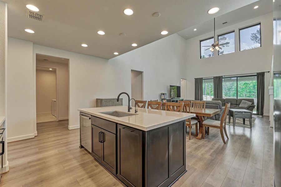 This modern open-concept living space features a spacious kitchen with a central island and sink, a dining area with a wooden table, and a cozy living room. Large windows and high ceilings create a bright, airy atmosphere, while the view of greenery adds a serene touch.
