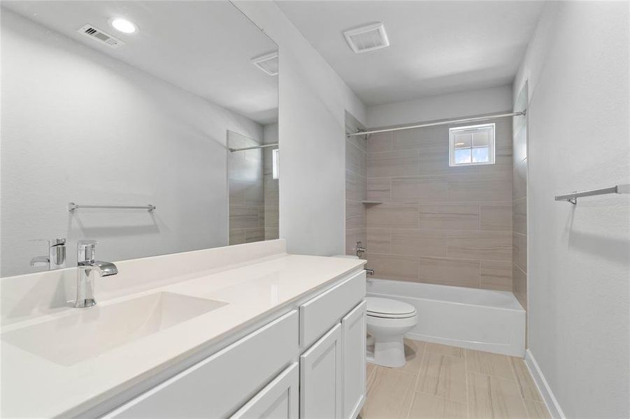 Secondary bath features tile flooring, bath/shower combo with tile surround, white stained wood cabinets, beautiful light countertops, mirror, dark, sleek fixtures and modern finishes.