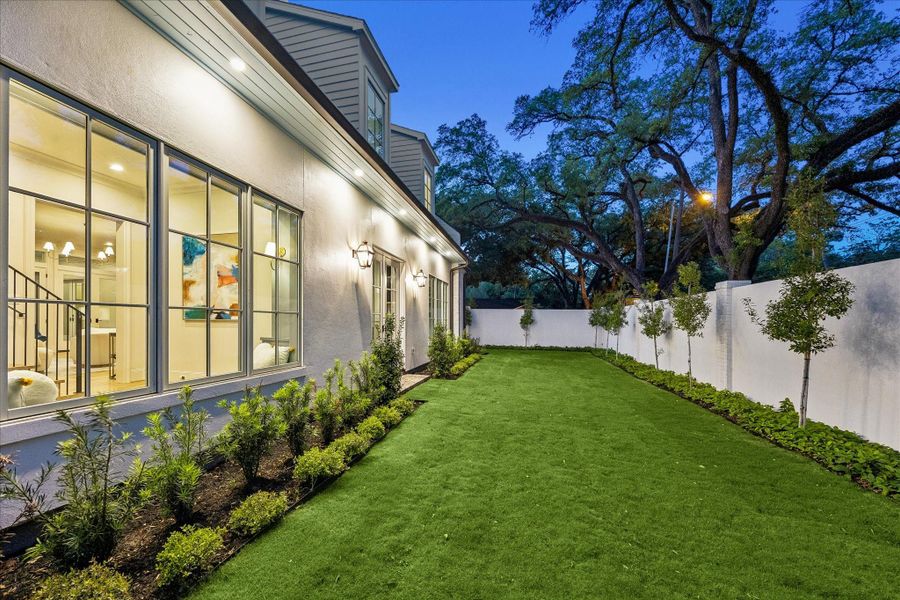 Another view of the landscaped grounds featuring lush turf, manicured landscaped beds & privacy wall. Large windows line the home, creating a seamless connection to the outdoor spaces while landscape lighting & trees enhance the setting.