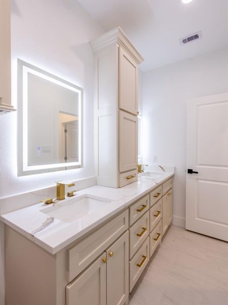 This bathroom features a modern design with dual sinks set in a sleek white vanity, accented by gold fixtures. The mirrored lighting adds elegance, and ample cabinetry provides plenty of storage.