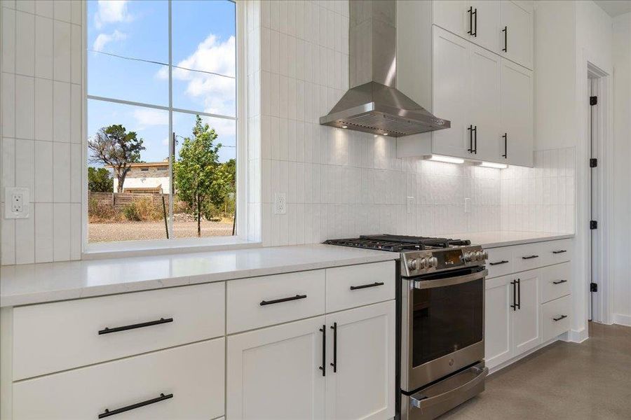 Kitchen with wall chimney range hood, stainless steel range with gas stovetop, white cabinetry, decorative backsplash, and concrete floors