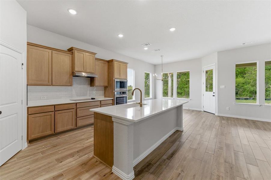 Kitchen with light wood-type flooring, a kitchen island with sink, decorative backsplash, stainless steel appliances, and light stone counters