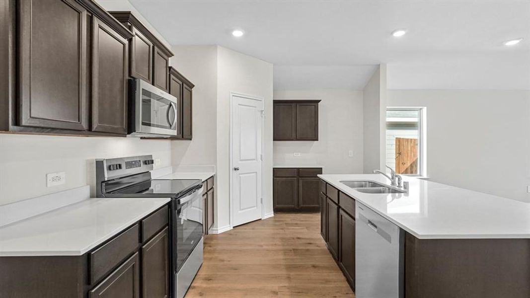 Kitchen featuring dark wood finish cabinetry, stainless steel appliances, light wood-type flooring, a center island with sink, and recessed lighting