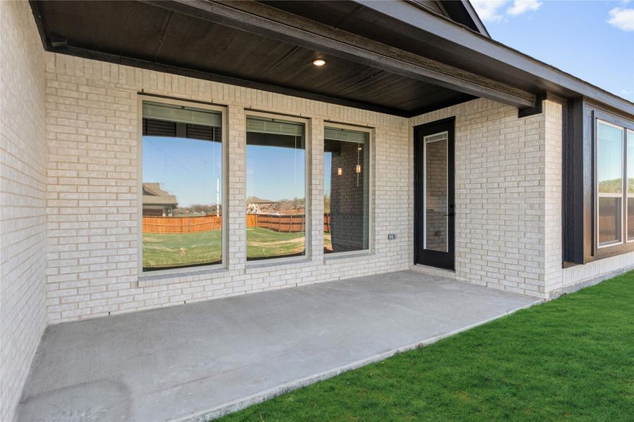 Covered back patio with concrete slab, white brick exterior, large windows, and black door overlooking green lawn Covered back patio with concrete slab, white brick exterior, large windows, and black door overlooking green lawn