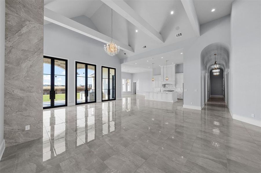 Unfurnished living room with a chandelier, high vaulted ceiling, healthy amount of natural light, beam ceiling, and light marble finish floors Unfurnished living room with a chandelier, high vaulted ceiling, healthy amount of natural light, beam ceiling, and light marble finish floors