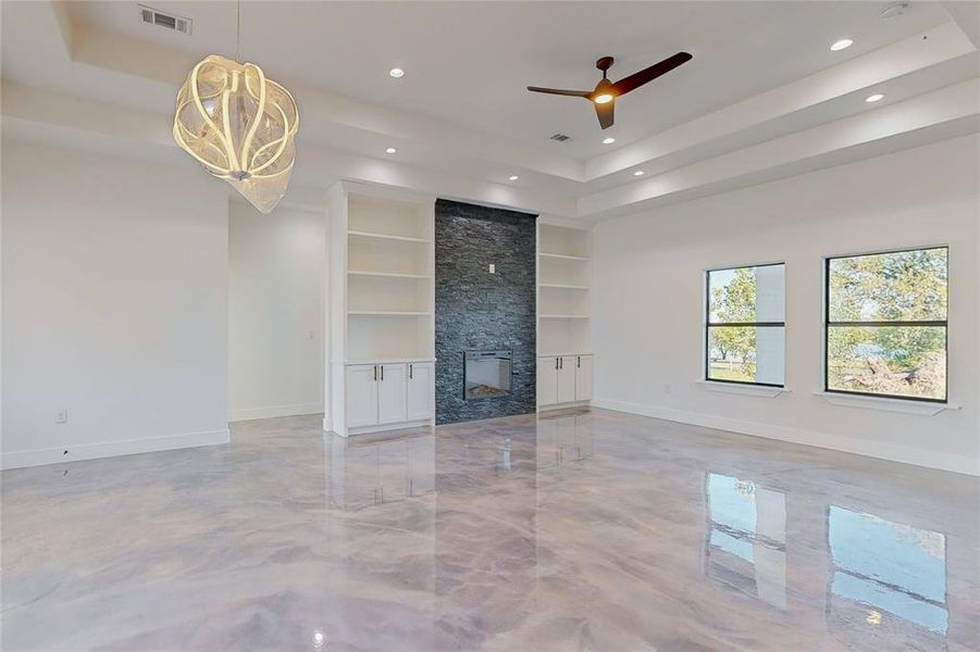 Unfurnished living room with built in features, ceiling fan with notable chandelier, a stone fireplace, and a tray ceiling