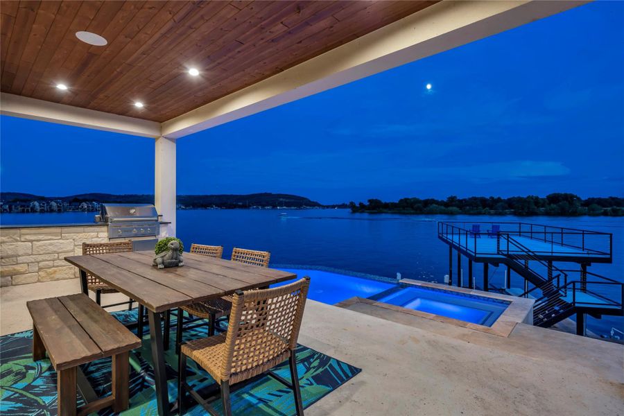 Patio at night with a water view, outdoor dining area, a patio area, a pool with connected hot tub, and area for grilling