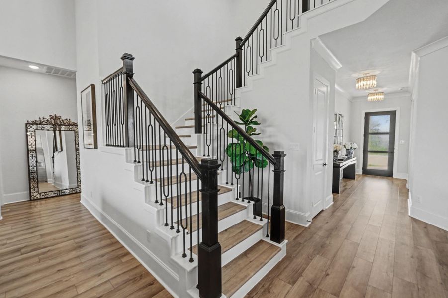 A striking staircase with dark wood handrails and detailed iron spindles anchors the main entry hall. The light wood-look floors lead toward a welcoming front door and an elegant crystal-accented light fixture hangs in the adjacent corridor.