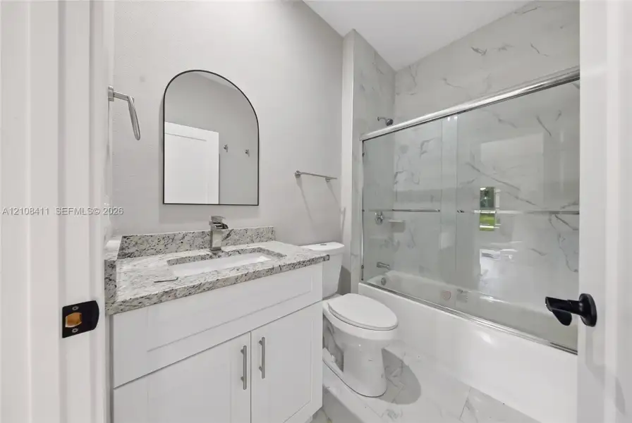 Bathroom featuring a white shaker-style vanity, stone-finish countertop, arched wall-mounted mirror, and a shower-tub combination with full-height tile surround