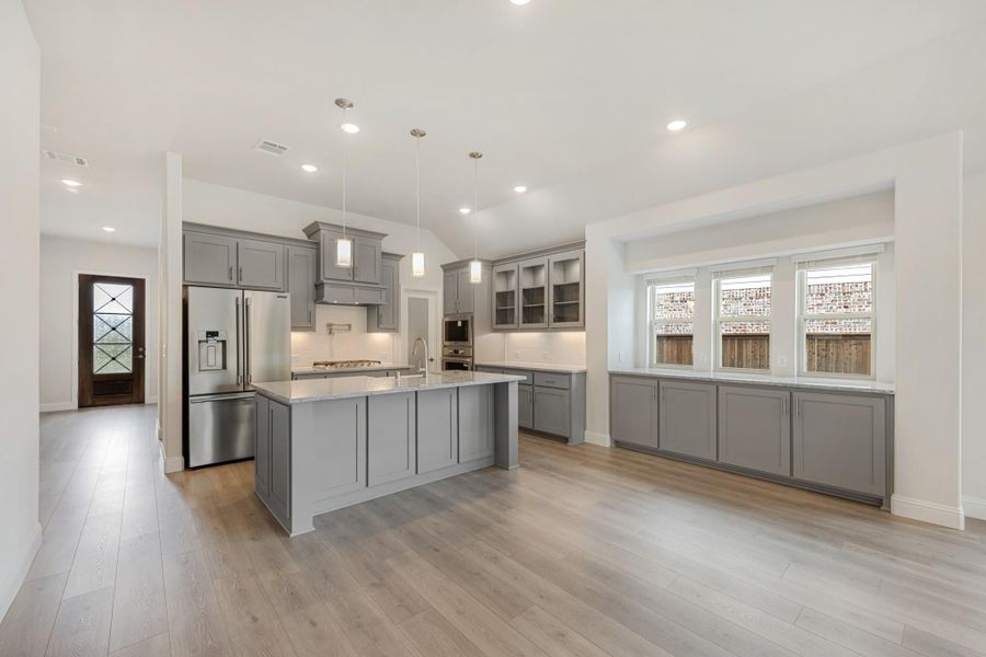 Open kitchen with gray cabinets, large island, stainless steel refrigerator, and light hardwood floors Open kitchen with gray cabinets, large island, stainless steel refrigerator, and light hardwood floors