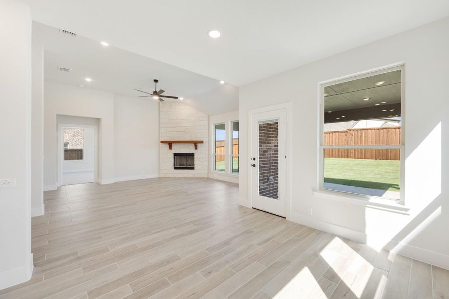 Open living room with light wood-look tile floors, stone fireplace, ceiling fan, and backyard views through large windows