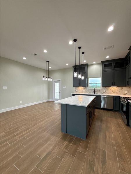 This is a modern kitchen with high ceilings and recessed lighting. It features dark cabinetry, white marble countertops, a central island, and stainless-steel appliances. The open layout and wood-look tile flooring enhance the spacious feel.