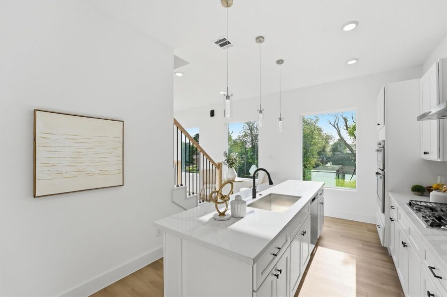 Bright, open kitchen featuring a spacious quartz island with waterfall edge, white shaker cabinetry, pendant lighting, wood-look flooring, and abundant natural light with views of surrounding greenery.