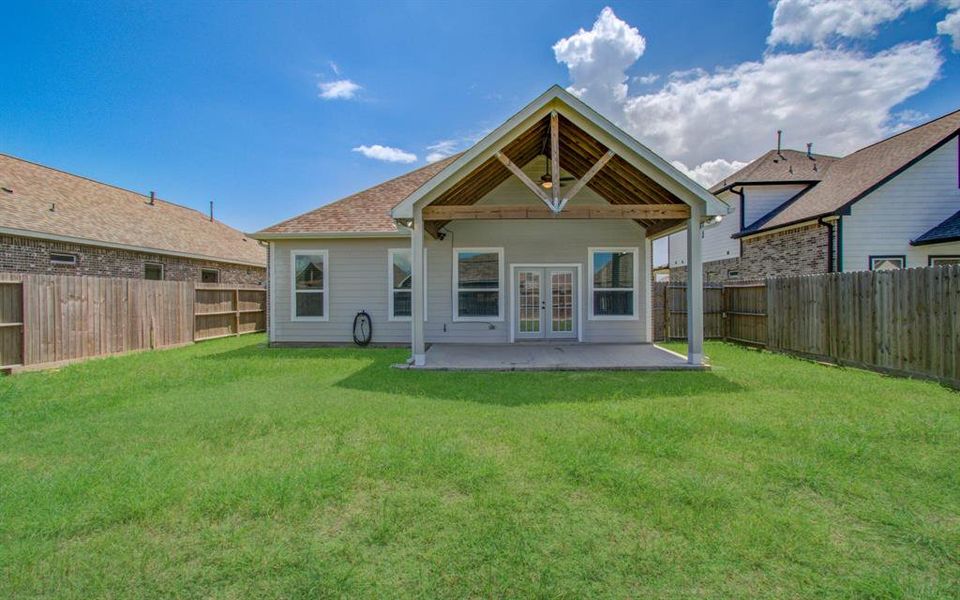 This photo features the rear exterior of a single-story home showcasing a covered patio with a pitched roof and ceiling fan, large windows, and a spacious, fenced-in backyard with freshly cut grass.