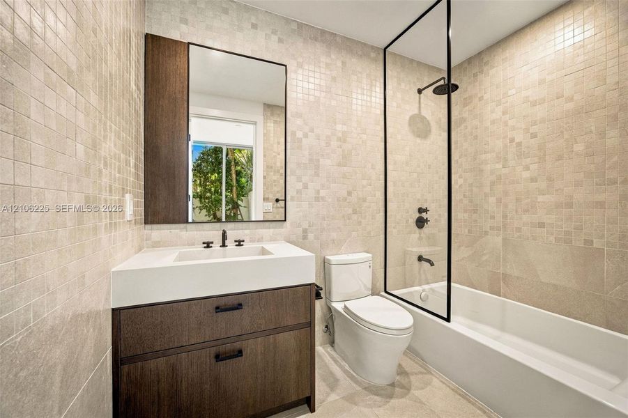 Bathroom featuring a wood-finish vanity with a rectangular sink, full-wall mosaic tiling, a glass-enclosed shower with matte black fixtures, and a wall-mounted mirror Bathroom featuring a wood-finish vanity with a rectangular sink, full-wall mosaic tiling, a glass-enclosed shower with matte black fixtures, and a wall-mounted mirror