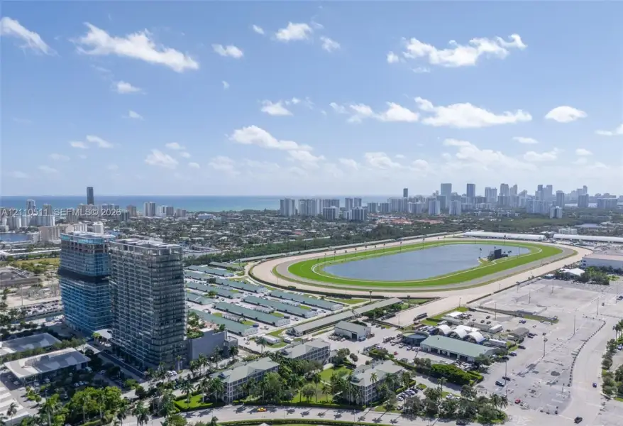 Aerial perspective featuring a multi-story glass-facade residential building, a large-scale race track, and a coastal skyline backdrop