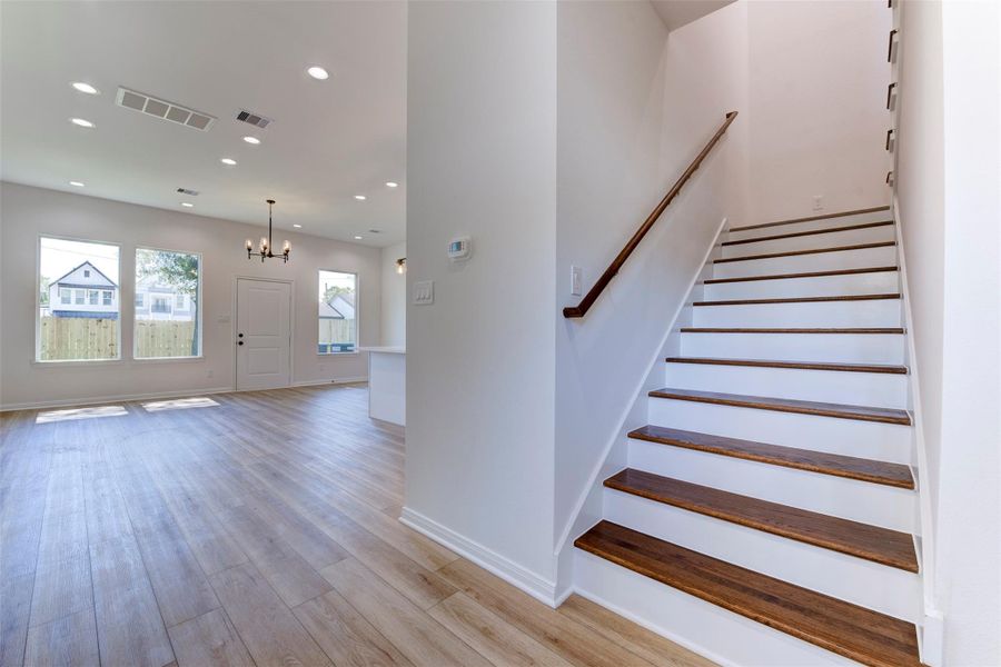 Seamless transition into the open-concept living space highlighted by wood-look LVP flooring, tall ceilings, and abundant natural light. Recessed lighting is thoughtfully layered throughout. Staircase features warm wood treads with crisp white risers and a clean handrail, adding a refined contrast to the home’s bright, neutral palette.