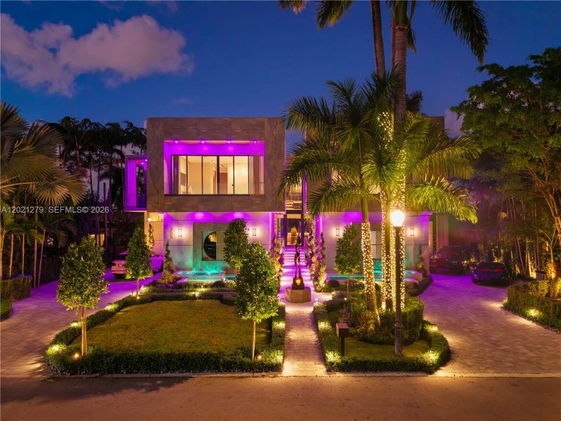 Front elevation at night showcasing stone facade, paver entry walkway, bronze sculpture, manicured hedges, motor court, illuminated reflecting pools, string-lit palms, and dramatic programmable violet LED uplighting across the full facade.