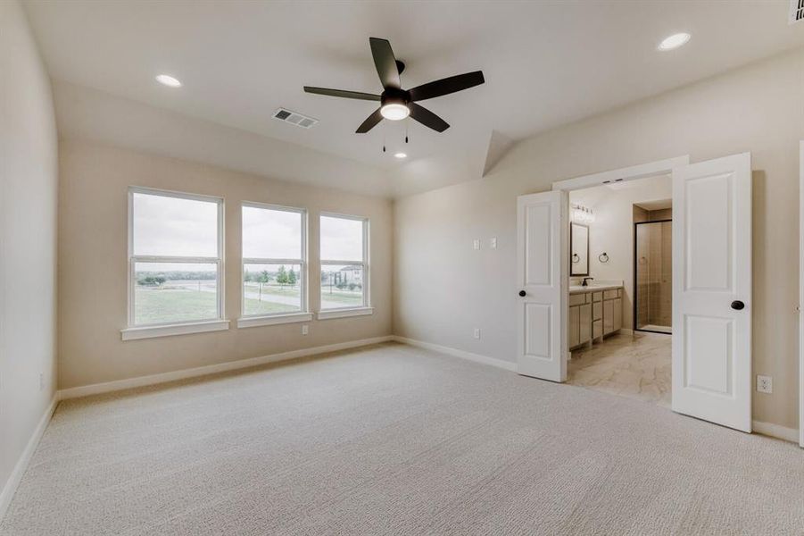 Spacious carpeted room featuring a vaulted ceiling with a ceiling fan, three large windows, recessed lighting, and double doors opening to an ensuite bathroom with a double vanity and framed glass shower