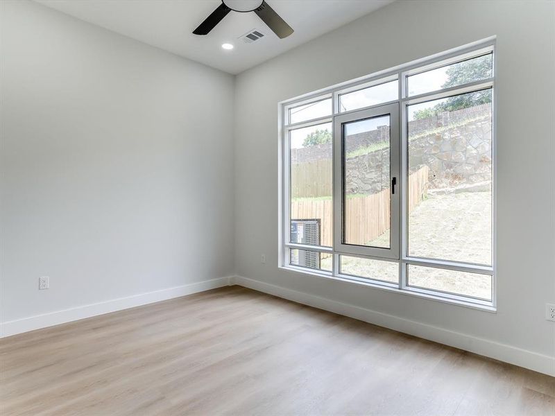 Downstairs guest bedroom. Spacious interior featuring a large multi-pane window, light wood-finish flooring, a contemporary ceiling fan, recessed lighting, and crisp white baseboards Downstairs guest bedroom. Spacious interior featuring a large multi-pane window, light wood-finish flooring, a contemporary ceiling fan, recessed lighting, and crisp white baseboards