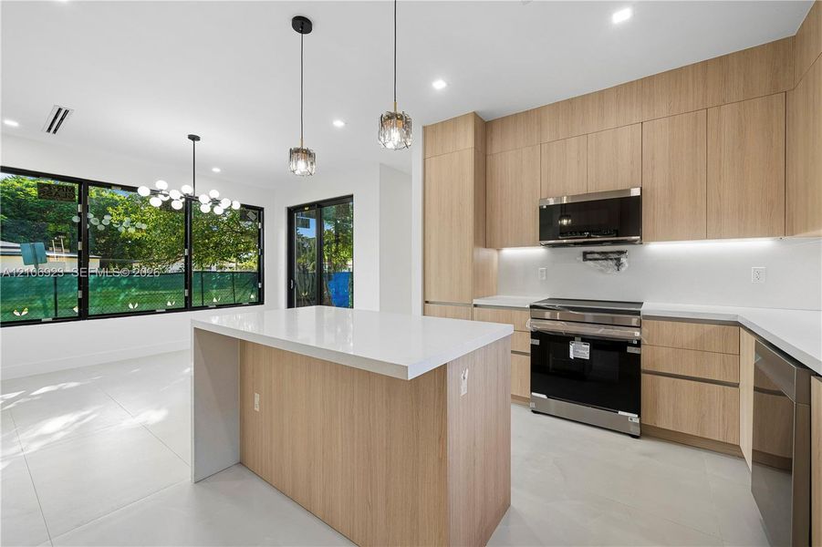 Kitchen featuring light wood-finish cabinetry, a central island with a white countertop, large-format tile flooring, stainless steel appliances, and integrated under-cabinet lighting Kitchen featuring light wood-finish cabinetry, a central island with a white countertop, large-format tile flooring, stainless steel appliances, and integrated under-cabinet lighting