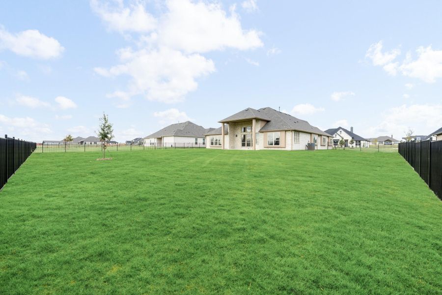 Large backyard with green lawn, black metal fence, and stone home with covered patio in background Large backyard with green lawn, black metal fence, and stone home with covered patio in background
