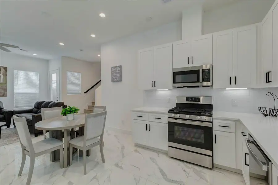 Open kitchen and dining view with stainless gas range, built-in microwave, dishwasher, white cabinetry, under-cabinet lighting, and a round dining area nearby. The layout connects easily to the living room and staircase, creating a bright, efficient main-level flow.
