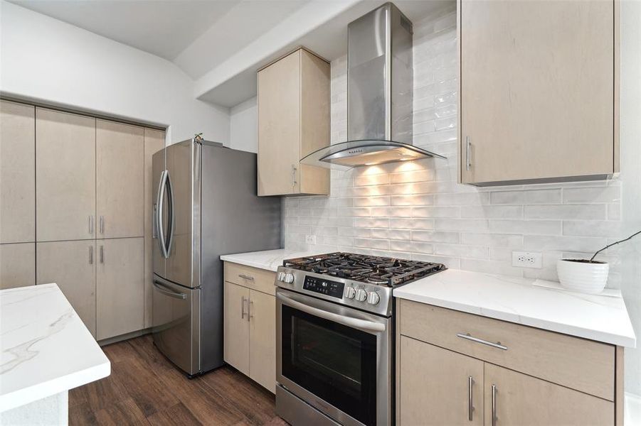 Kitchen featuring light brown cabinetry, tasteful backsplash, stainless steel appliances, dark wood-type flooring, and wall chimney range hood