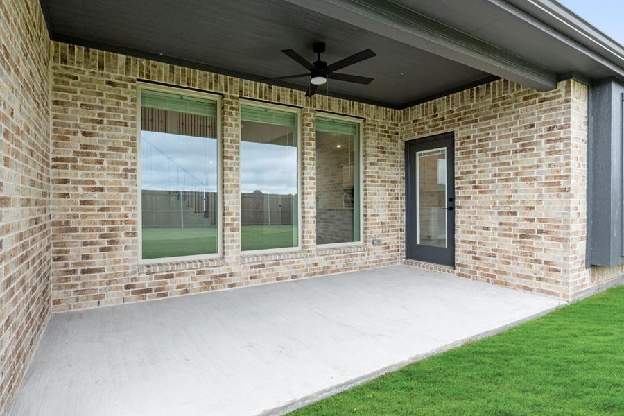 Covered back patio with ceiling fan, brick exterior, large windows, and concrete floor next to green lawn