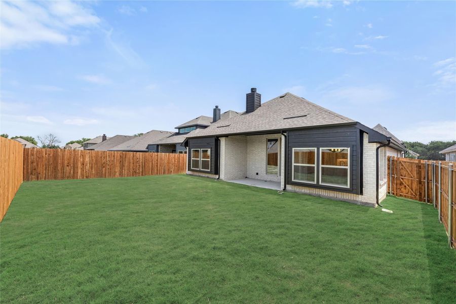Backyard of single-story brick home with dark trim, covered patio, green lawn, and wood privacy fence.