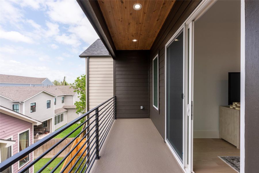 A second look at the balcony: continuous cedar ceiling with downlights, dark stained vertical board-and-batten accent, horizontal slat metal guardrail, full-height slider at the bedroom, sealed deck surface, and western exposure for afternoon shade.