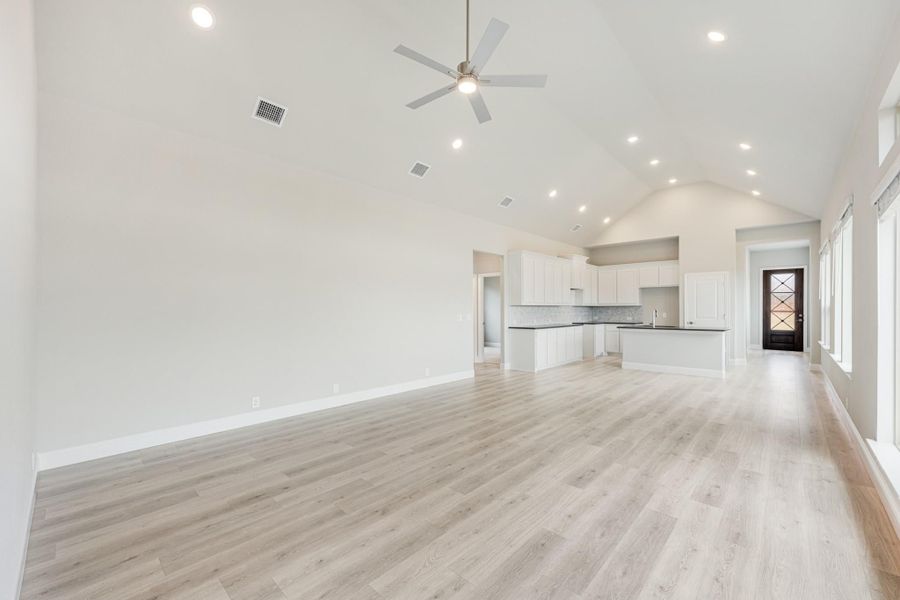 Open living area with vaulted ceiling, ceiling fan, light wood floors, and open kitchen with white cabinets in background