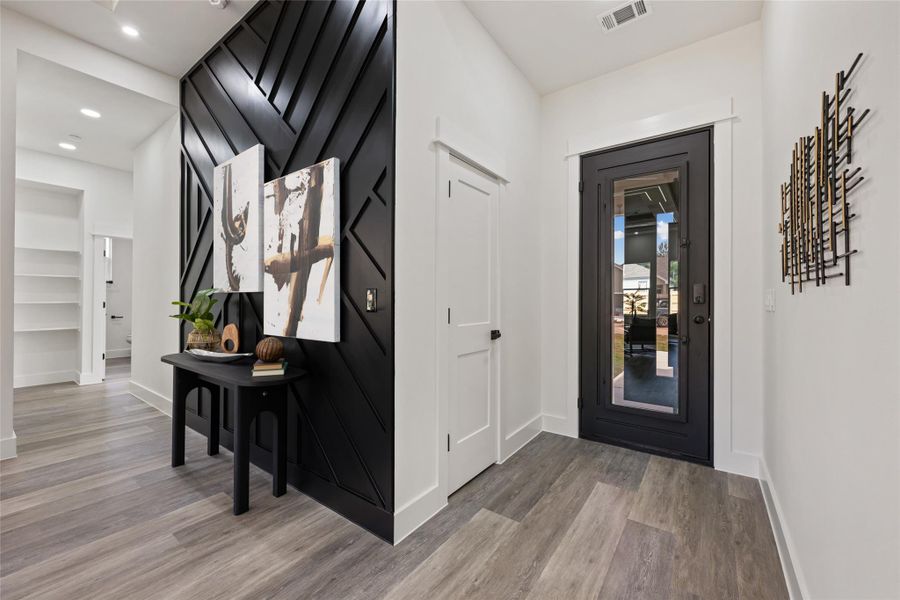 Entryway featuring a striking black accent wall with geometric paneling, wood-finish flooring, a modern black paneled door with glass insert, recessed lighting, and white baseboards