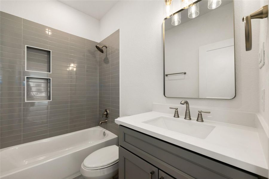 This secondary bathroom boasts tile flooring, a bath/shower combo with a stylish tile surround, stained wood cabinetry, elegant light countertops, a mirror, and sleek fixtures complemented by modern finishes.