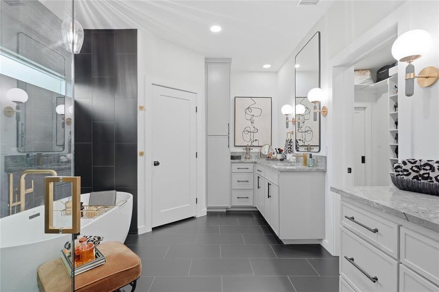 Freestanding soaking tub with gold-tone fixtures, enclosed glass shower, dual vanity with quartz countertops, custom cabinetry, and dark tile flooring